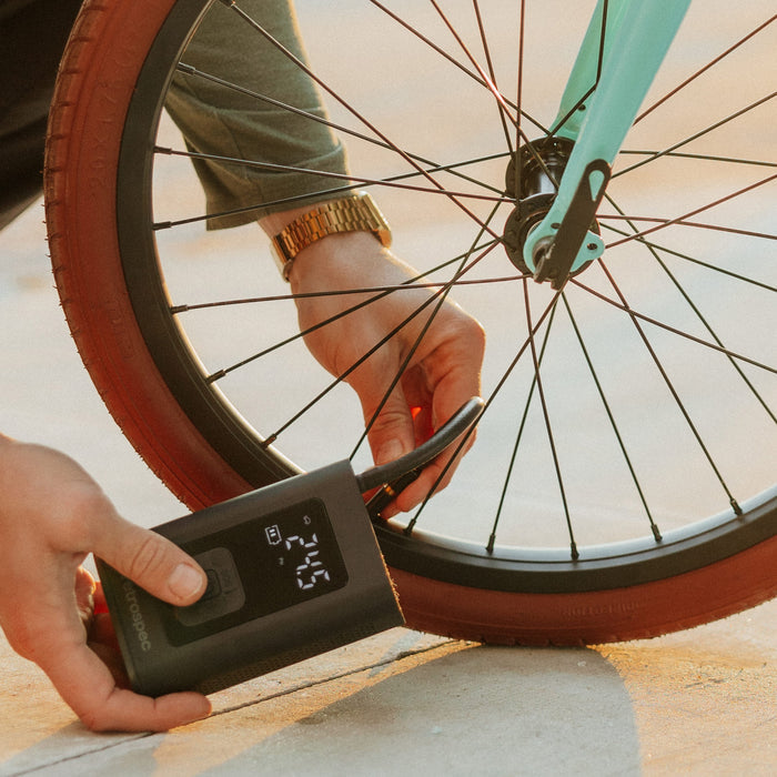 A person uses a Aer Portable Air Compressor, a portable air pump to inflate a bike tire, displaying a digital pressure gauge on the device as the sun sets.