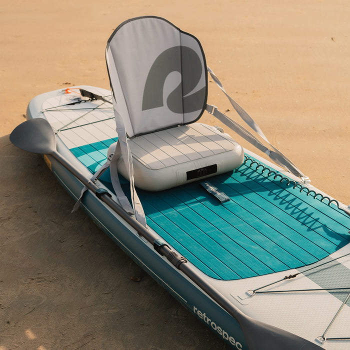 Inflatable stand-up paddleboard with an attached inflatable seat on a sandy beach.