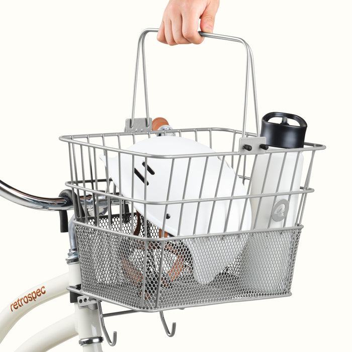 A silver bicycle basket with a white bottle and black container attached to a bike, held by a hand.
