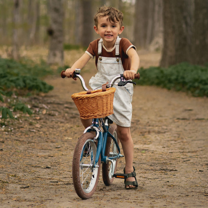 A young child in overalls rides a blue Beaumont Mini 16" Kids' Bike with a woven basket down a tree-lined path in a forested area.