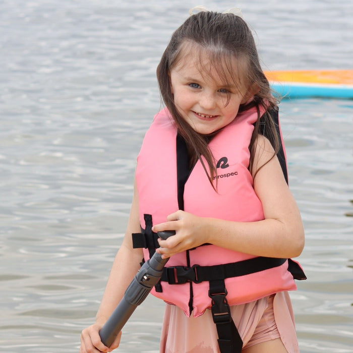 A smiling young girl wearing a retrospec Bobbin’ Child Life Jacket in Pink Lemonade stands at the water's edge holding a paddle, with a colorful Paddle Board visible in the background.