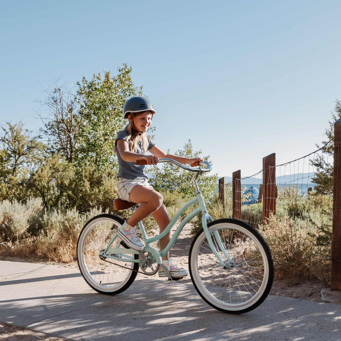 Child riding a light blue bicycle on a sunny day with trees and a fence in the background.