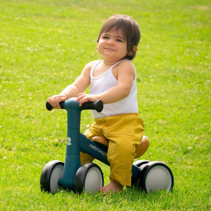 A young child sits on a blue Cricket 2 Baby Walker Balance Bike, wearing a white tank top and yellow pants, surrounded by lush green grass.