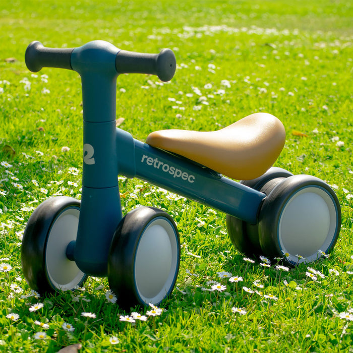 A blue retro-styled Cricket 2 Baby Walker Balance Bike with a tan seat, resting on green grass sprinkled with white daisies, showcasing its playful design.