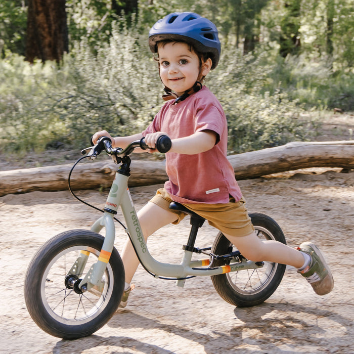 A child rides a Cub Plus 12" Kids' Balance Bike through a forested path, wearing a helmet and casual clothing, enjoying outdoor play in nature.