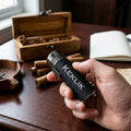 Hand holding black cigar lighter with monogrammed "KFLK" initials, torch lighter with metal band, wooden cigar box and cigars visible on desk background