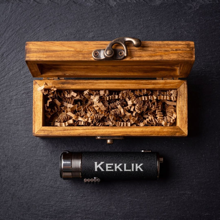 Open wooden box with shredded packing material revealing black cylindrical lighter engraved with sample text "KEKLIK" and bronze metal accents