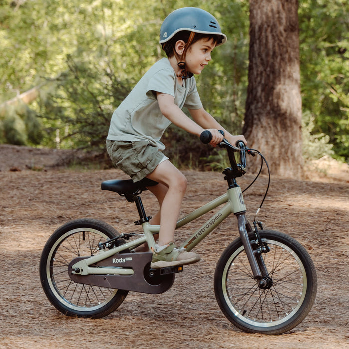 A child rides a Koda Plus 16" Kids' Bike on a wooded path, wearing a helmet, a light shirt, and shorts, surrounded by trees and pine needles.