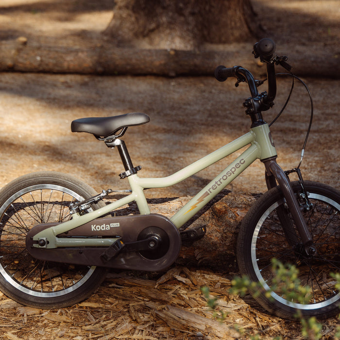 A light green retro-style Koda Plus 16" Kids' Bike leans against a log in a natural setting, showcasing its sleek frame and minimalistic design.