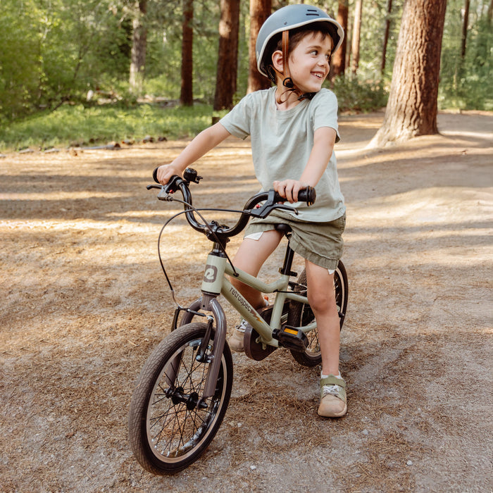 A child in a helmet rides a green Koda Plus 16" Kids' Bike on a dirt path surrounded by tall trees in a sunny, wooded area.