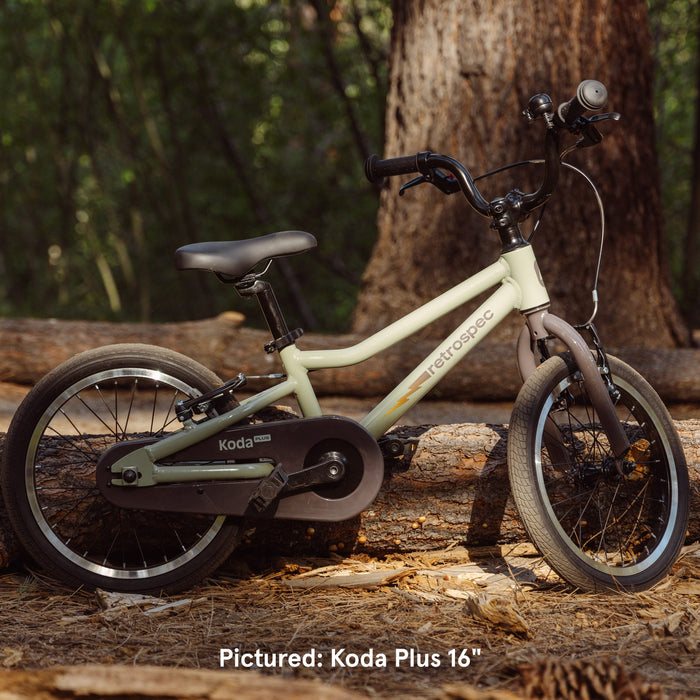 A Koda Plus 16" bike in soft green, resting against a log in a woodland setting, showcasing its sleek design and sturdy frame.