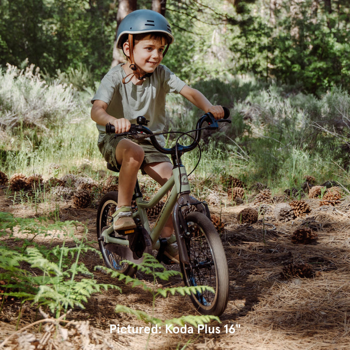A child rides a Koda Plus 16" bike on a forest trail surrounded by pinecones and greenery. The scene is sunlit and vibrant.