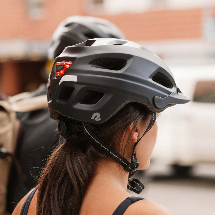 Rear three-quarter view of a woman wearing the retrospec Lennon Commuter Bike Helmet in matte black, showing the integrated red LED safety light, reflective strip, retrospec logo, and adjustable chin strap, with a second helmeted rider and urban buildings softly blurred behind her.