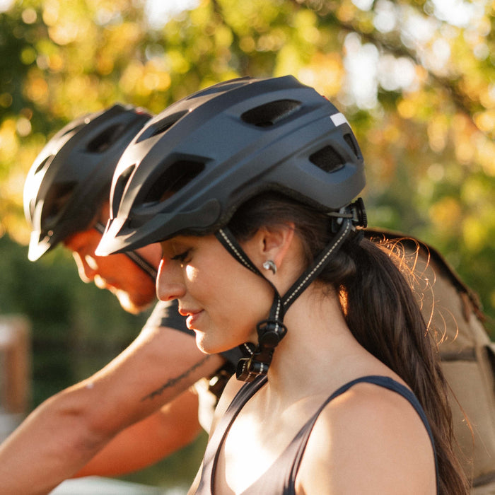Two people wearing their retrospec Lennon Commuter Bike Helmet in matte black at golden hour, with soft-focus autumn trees glowing in the background.