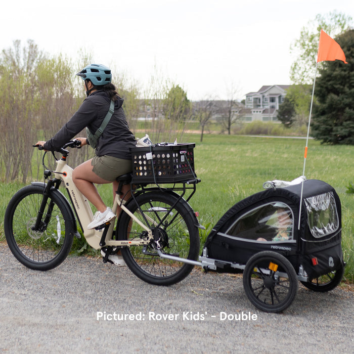 A woman rides an electric bike with a rear rack basket while towing a black Rover Kids' Bike Trailer on a gravel path.