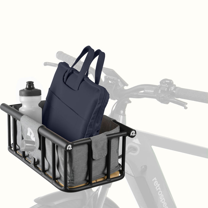 A metal ebike basket with a fabric gray liner holds a dark blue bag and a water bottle, with bike handlebars visible in the background.