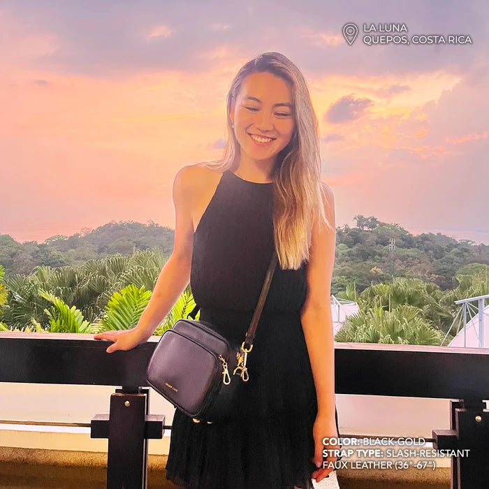 Woman in a black dress with a black handbag against a scenic background. Modeling the Verona Crossbody in black-gold with slash resistant faux leather strap