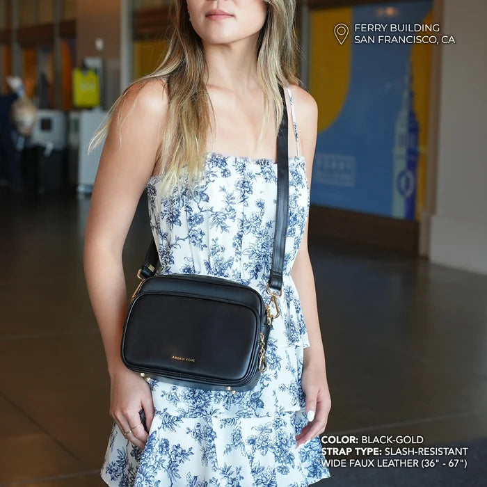 Woman holding a black handbag in an indoor setting. Modeling the Verona Crossbody in black-gold with slash resistant wide faux leather strap