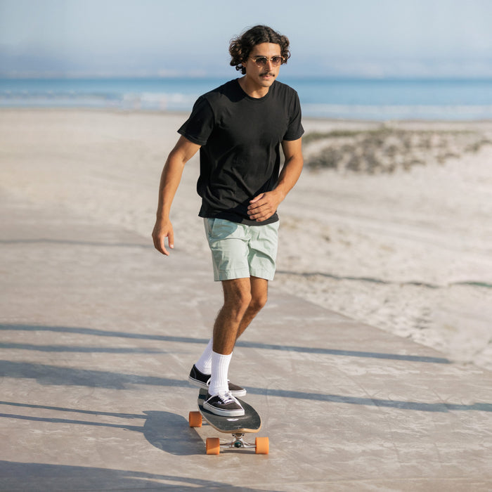 A person in a black t-shirt and green shorts skateboards along a sunny beach promenade, with the ocean visible in the background.