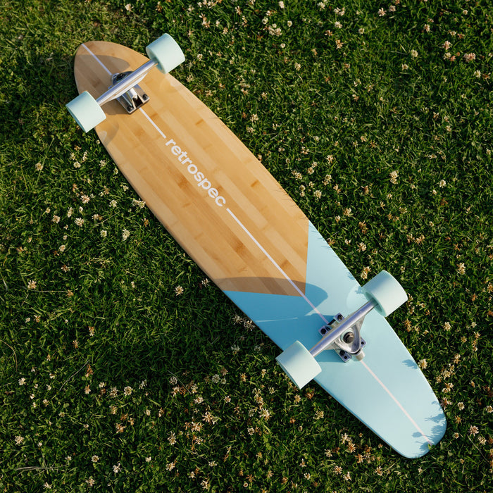 The Zed 44" Longboard skateboard with a bamboo deck and blue accents, resting on lush green grass sprinkled with tiny white flowers.
